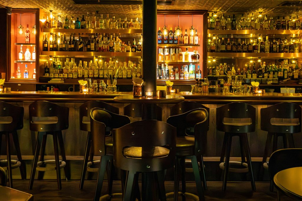 Balcão de bar em luz baixa, com banquetas altas e garrafas alinhadas nas prateleiras