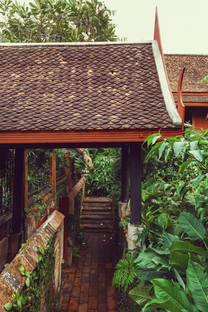Passagem de tijolos sob telhado de telhas de barro levando a um jardim tomado por trepadeiras