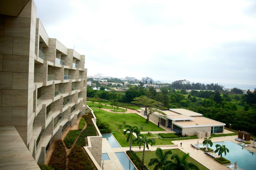 Fachada horizontal de hotel de linhas retas debrucado sobre jardim tropical e piscina, com a faixa do mar ao fundo