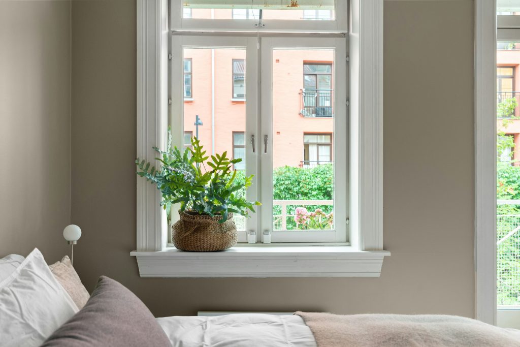 Cama encostada sob a janela com vasos de plantas no parapeito e vegetação verde do lado de fora