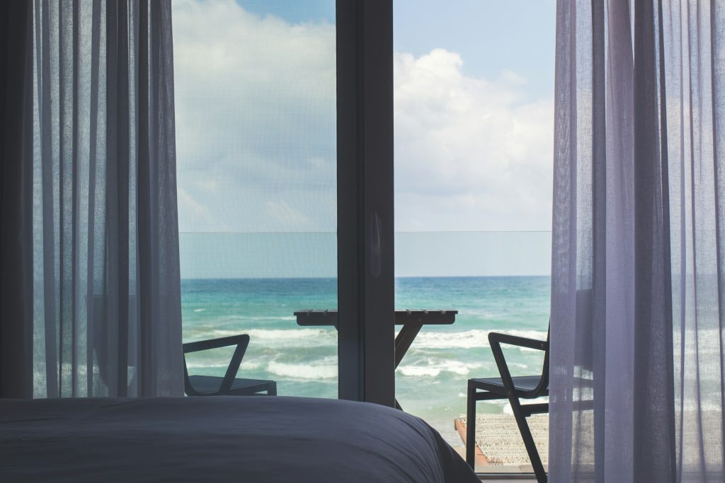 Interior de quarto de hotel com porta de vidro e cortina abertas para a varanda e o mar ao longe